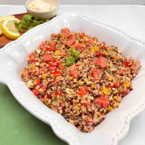 A white bowl filled with quinoa watermelon salad. A dish of feta cheese, mint and lemon slices are in the background cheese