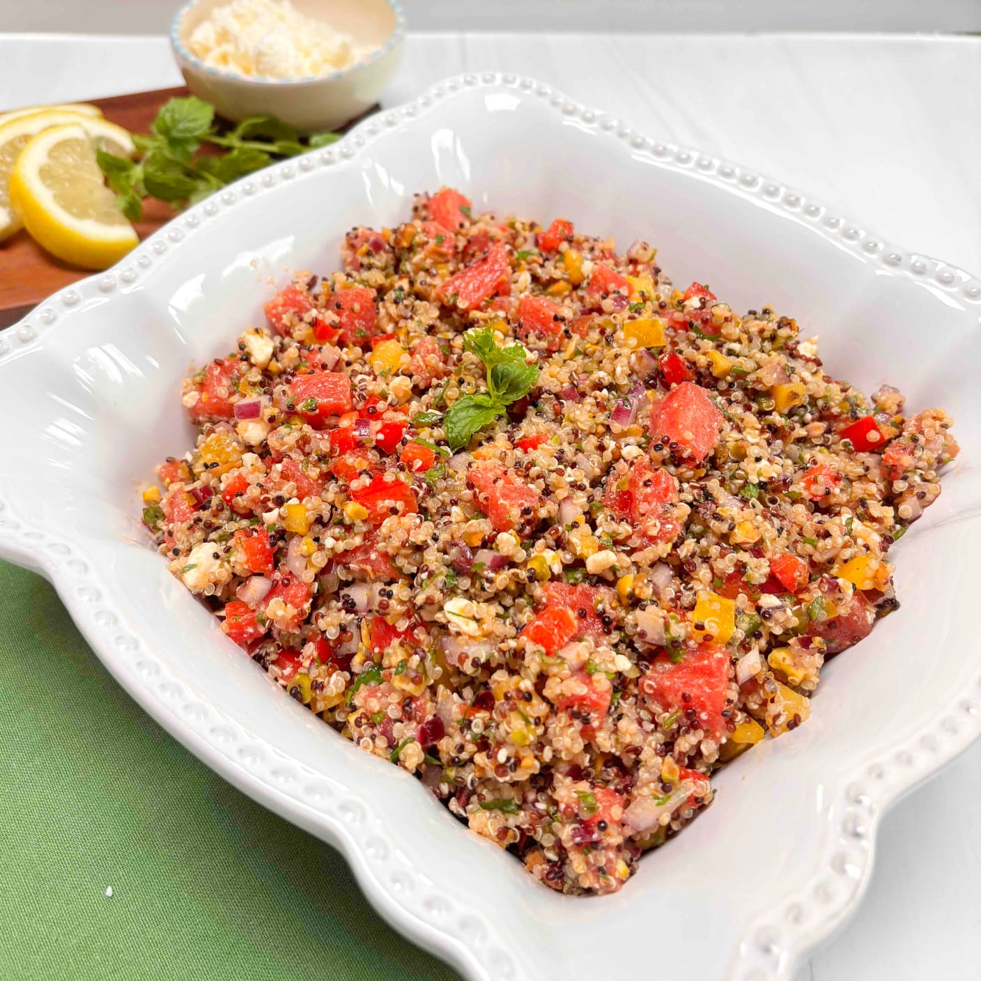 A white bowl filled with quinoa watermelon salad. A dish of feta cheese, mint and lemon slices are in the background cheese